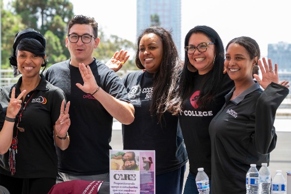 Six people at an information table wave into a camera