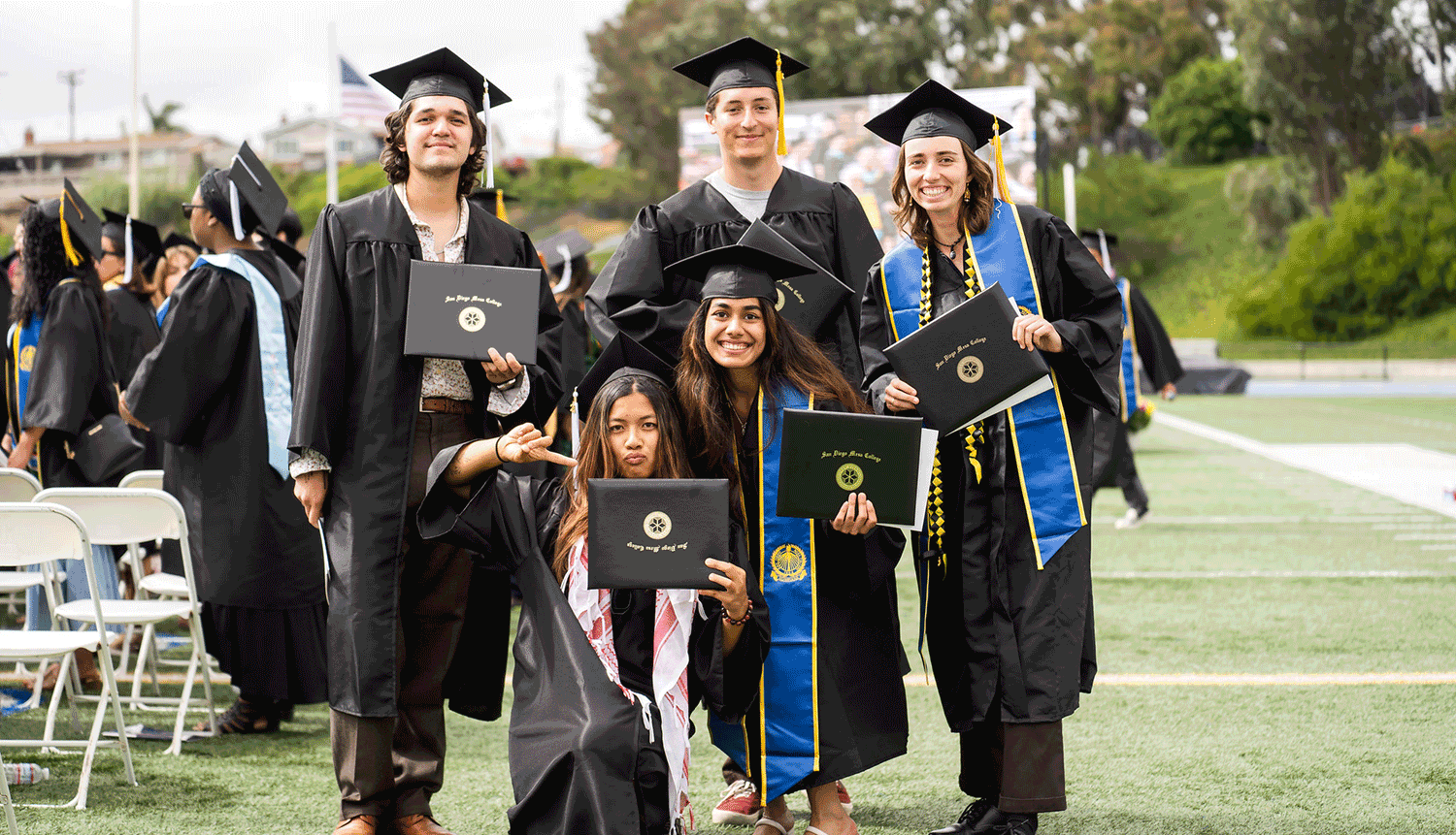 5 students in black graduation caps and gowns hold up their degrees.