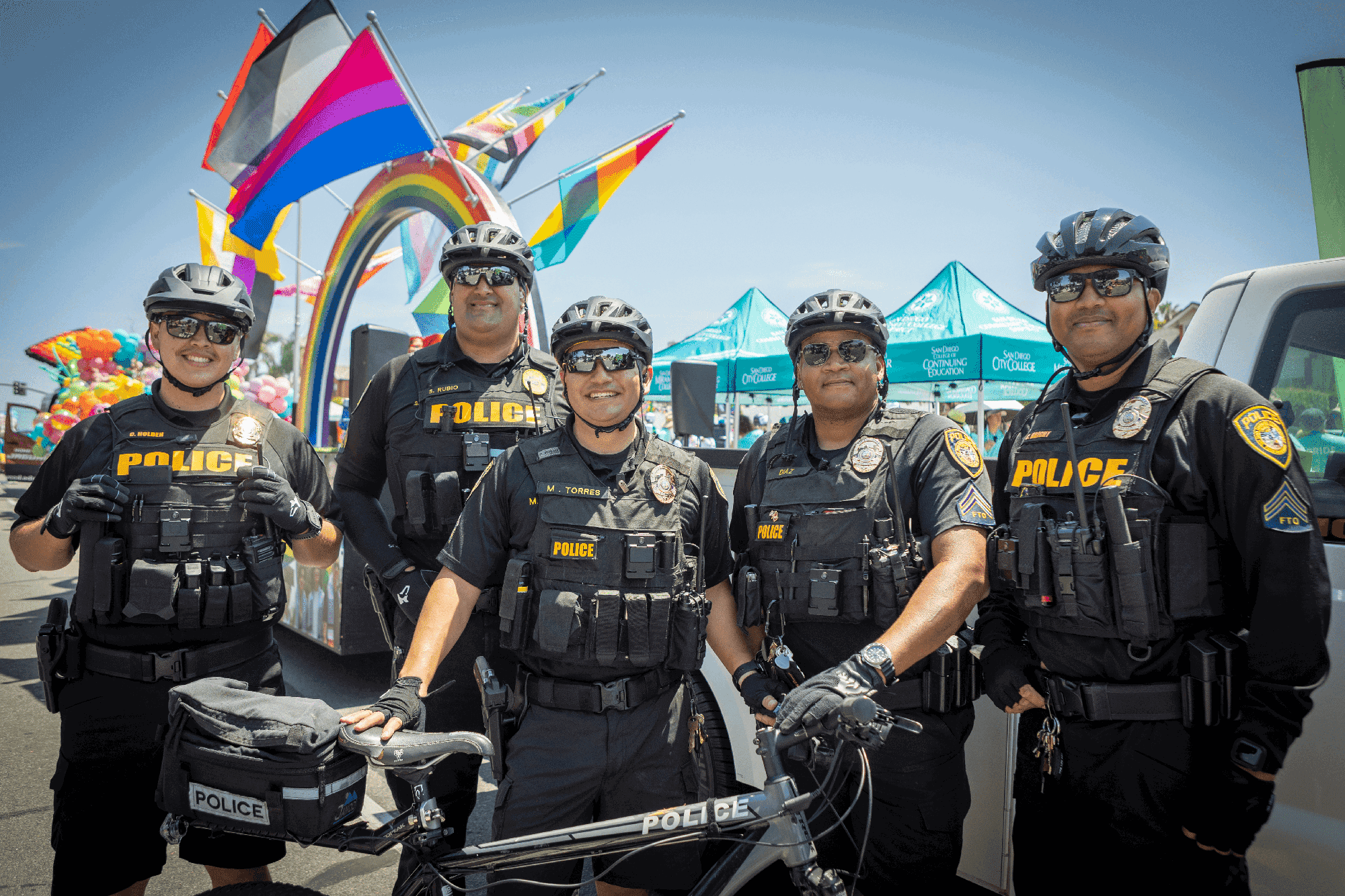Five college police wearing bike helmets and standing with a bicycle at the Pride Parade the pride flags are behind them