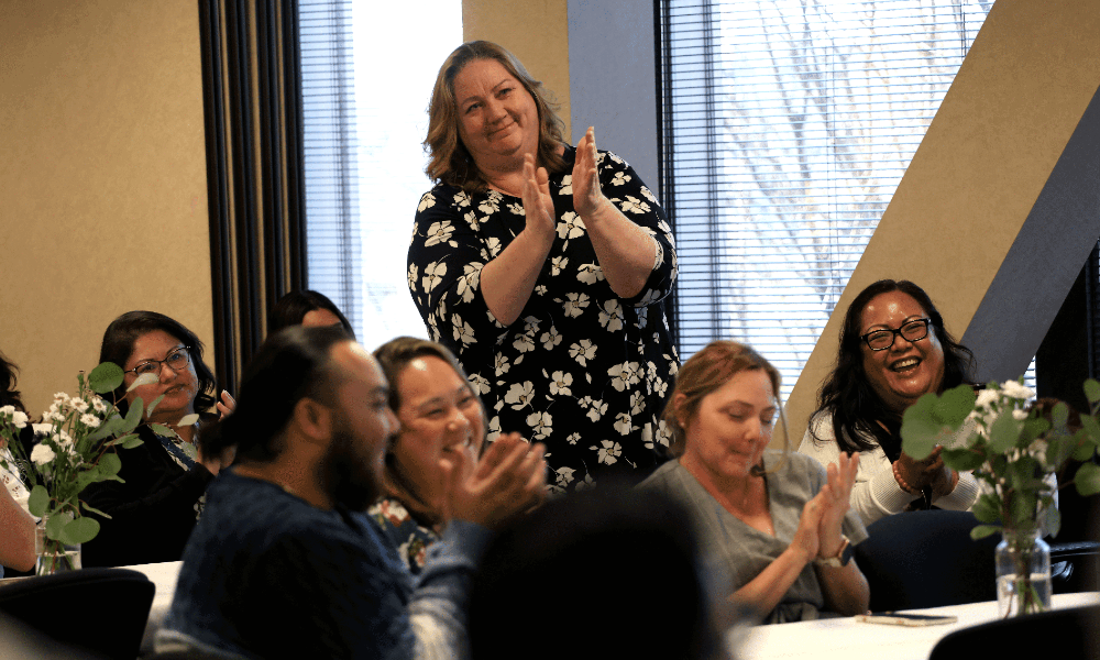 A woman in a room full employee stands and applauds at a reception