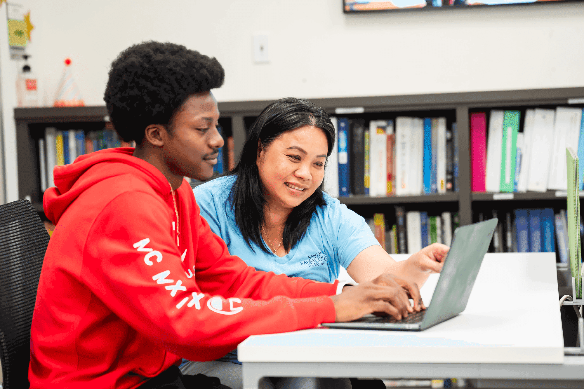 A student and an instructor sit at a table. The student is wearing a bright red sweatshirt and typing on a tablet