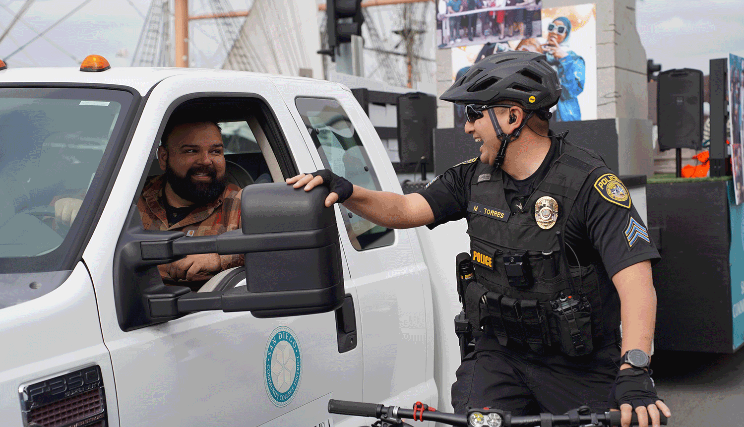 A College police officer rides his bike along side an SDCCD work truck pulling the float in the MLK parade. The driver and the officer are smiling and laughing.