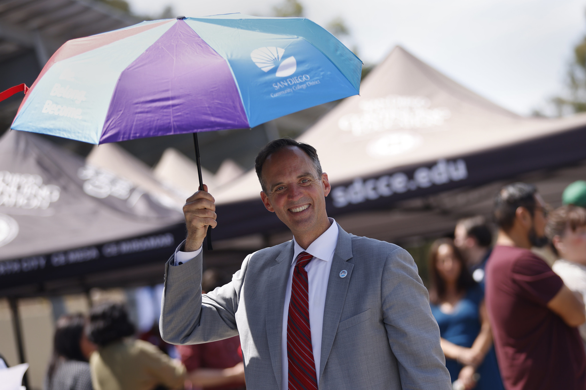 Greg Smith in a gray suit holds an umbrella with all the district colors.