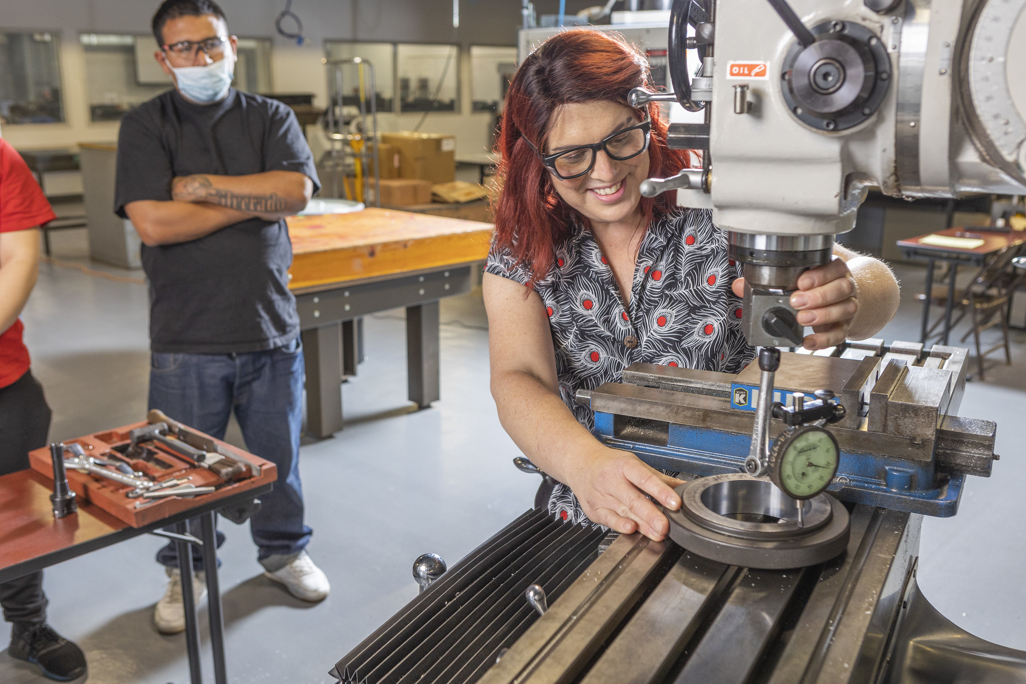 A student is a machine in a workshop class while another student looks on. looks on