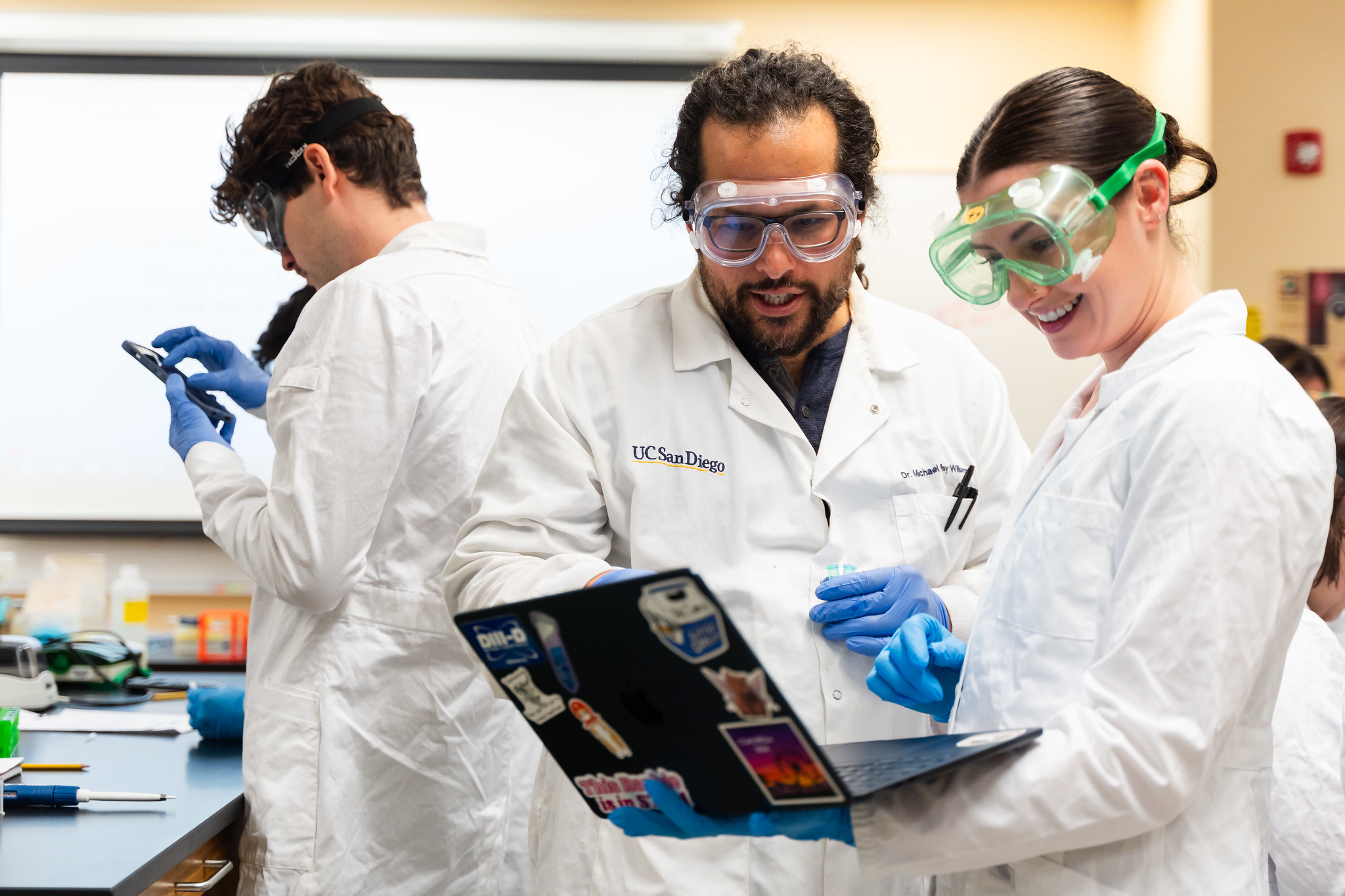 Two people in white lab coats and protective goggles looking at a laptop in a science lab.