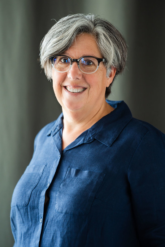 Lisa, a white woman with short gray hair and glasses, is wearing a dark blue shirt and smiles while looking at the camera