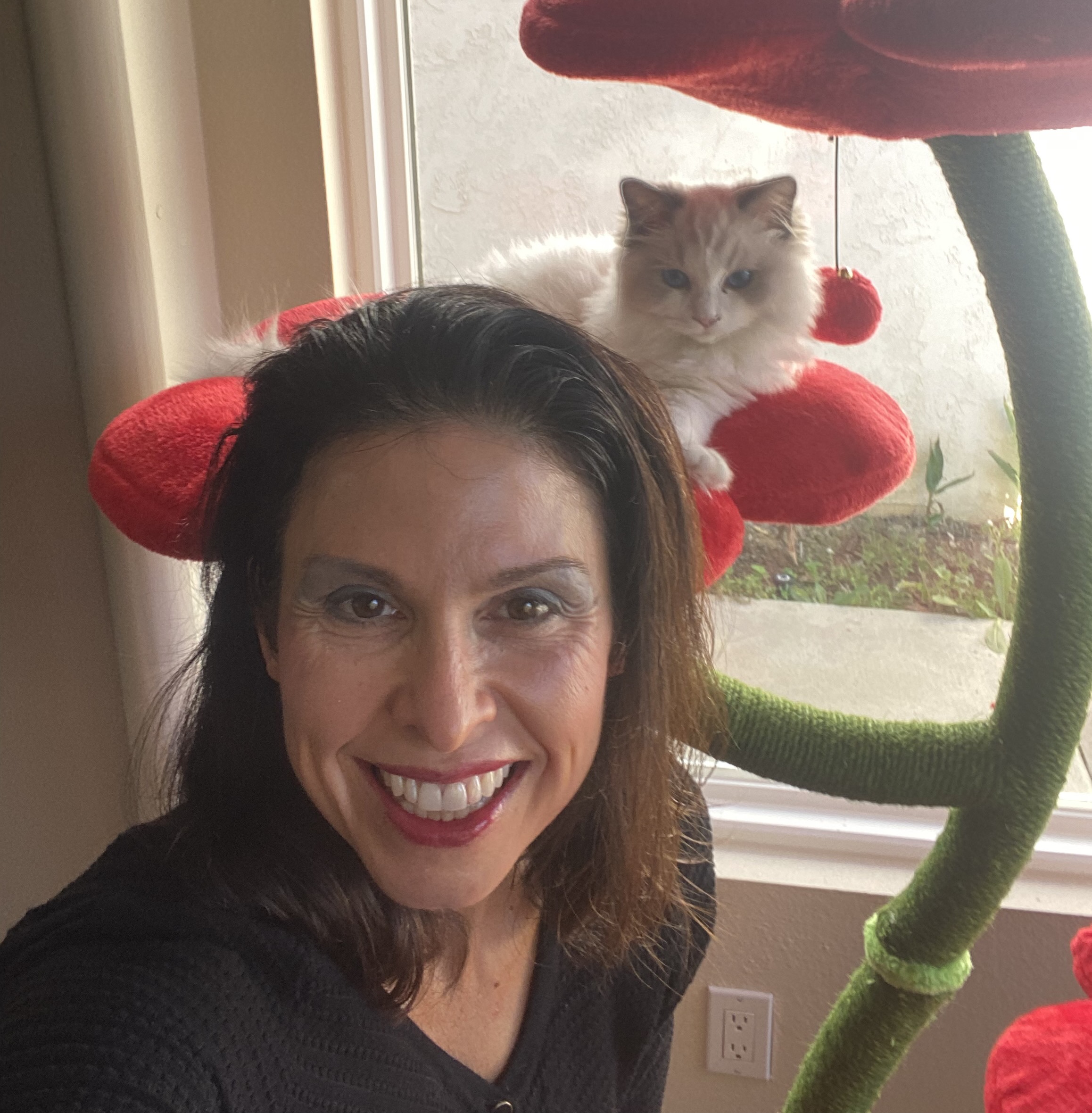 Melanie, an olive-skinned woman with brown hair, smiles at the camera wearing blue eyeshadow and red lipstick. Behind her is a white cat with long fur sat atop a red cat tree
