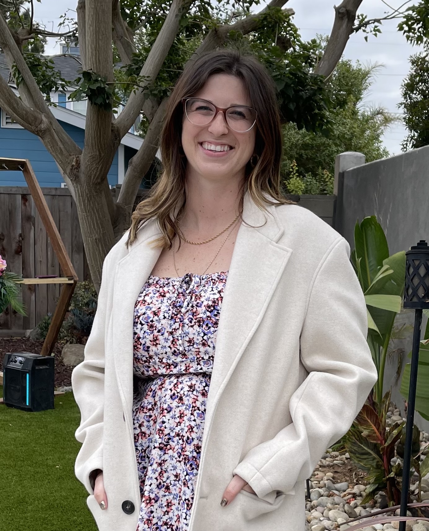 Maddie, a white woman with shoulder length brown hair and glasses, smiles at the camera, wearing a pink floral dress and white coat