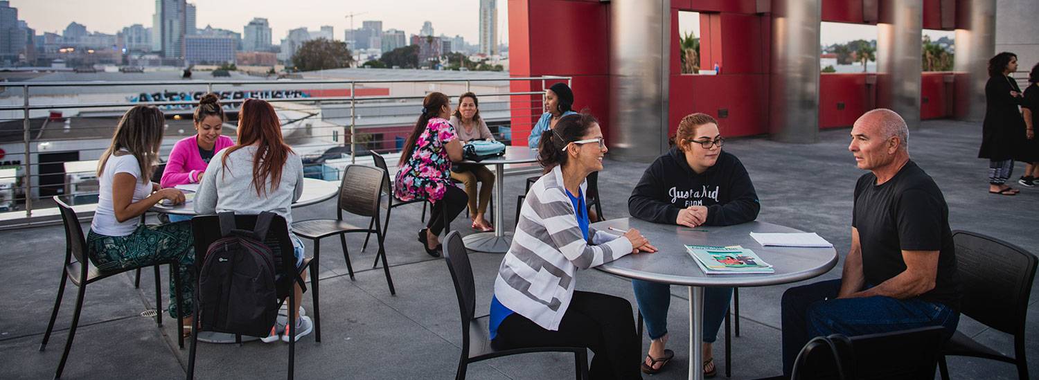 Eight people sit at tables on the rooftop of the Cesar Chavez campus. The San Diego skyline is in the background.