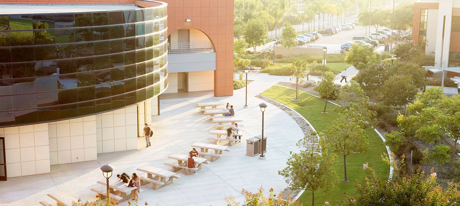 A birds eye view of a courtyard at Miramar College shows a building with students outside sitting on benches next to grass and a parking lot behind them.