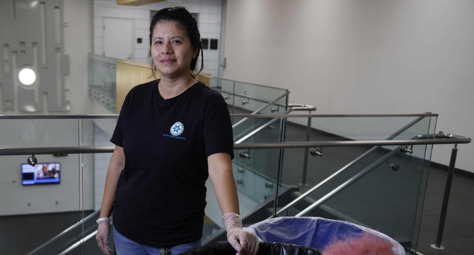 Edith Rangel in an upstairs hallway at the North City Campus. She is pushing a garbage can and cleaning supplies while wearing cleaning gloves.