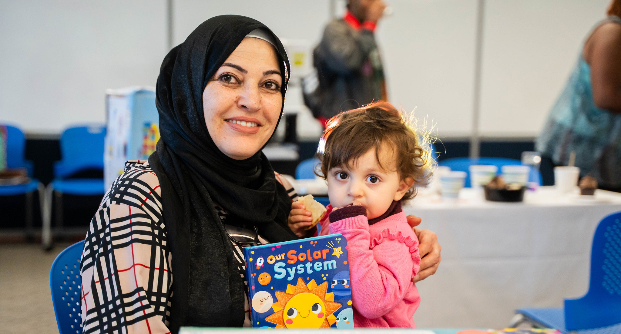 A woman sitting down at a table is holding a small child and a children's book