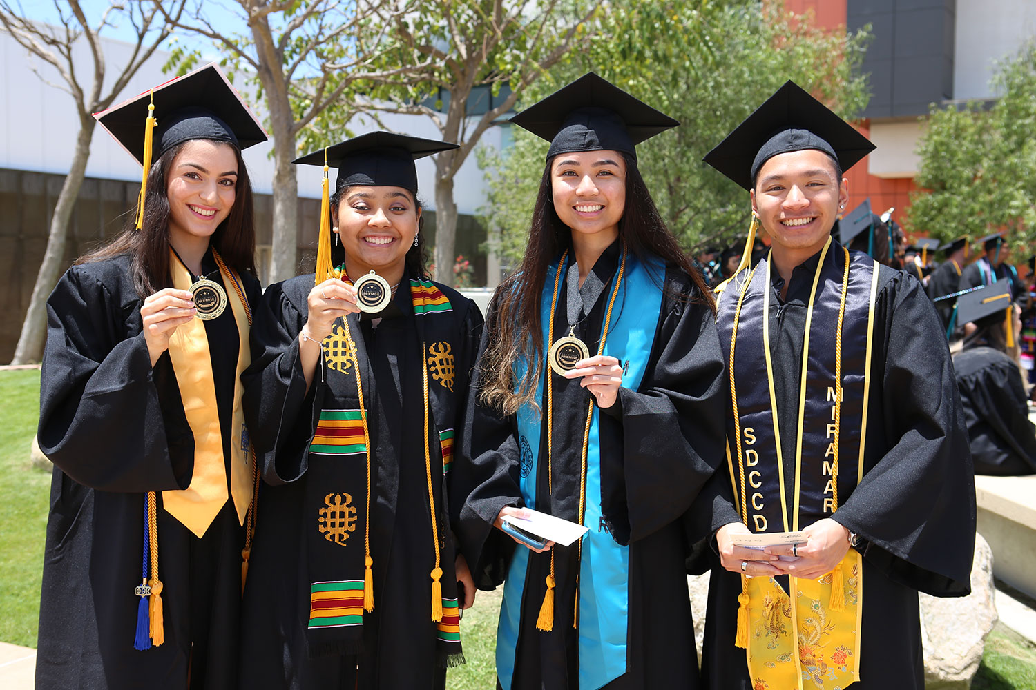 Four students in black graduation caps and gowns. Three hold up medallions for the promise program 