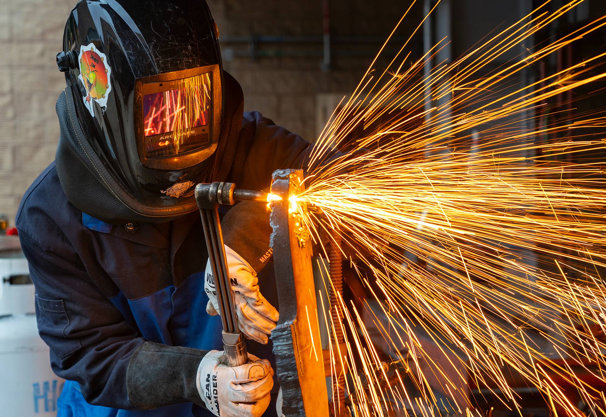 A welder in blue overalls, gloves and a welding mask uses a torch. Red sparks are flying around.
