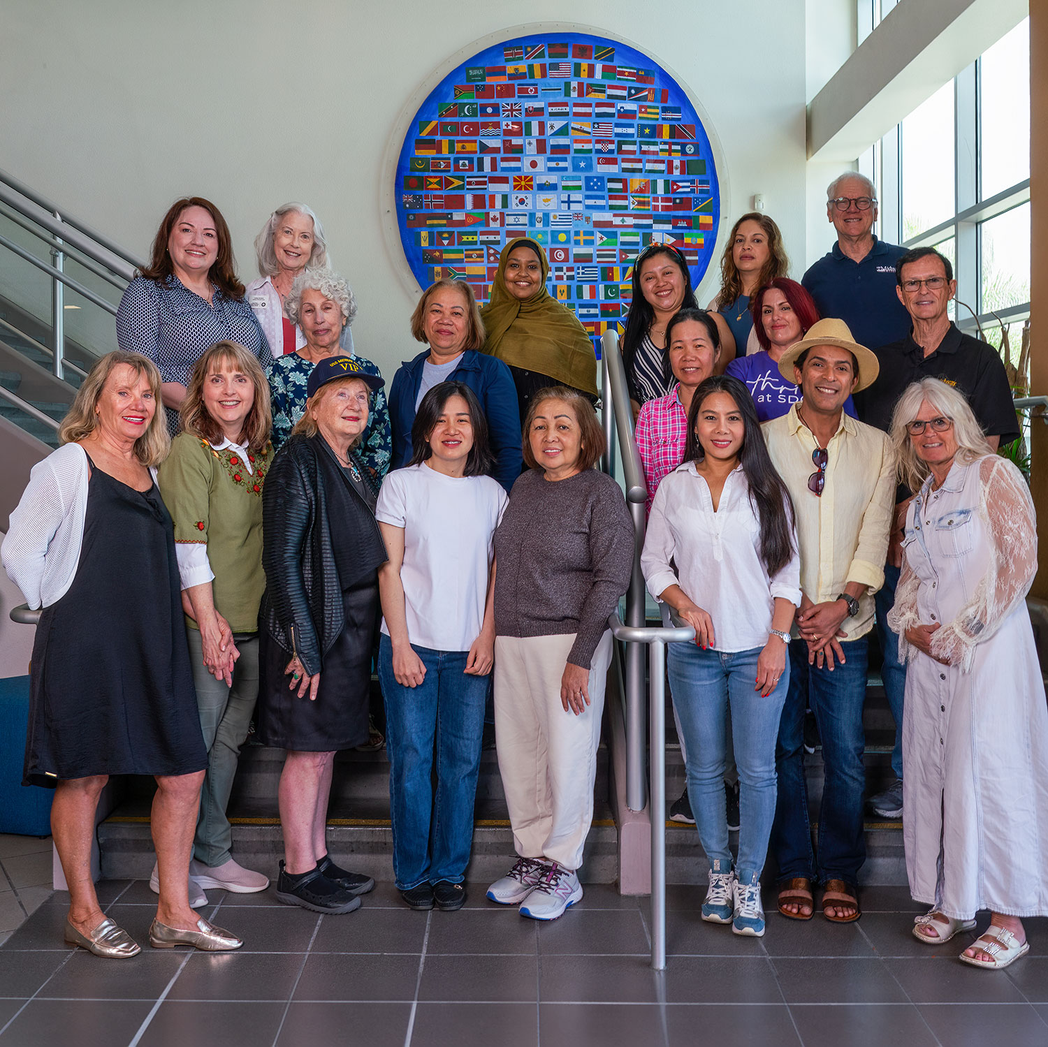 19 people gather on a staircase in front of a mural with flags from dozens of countries.