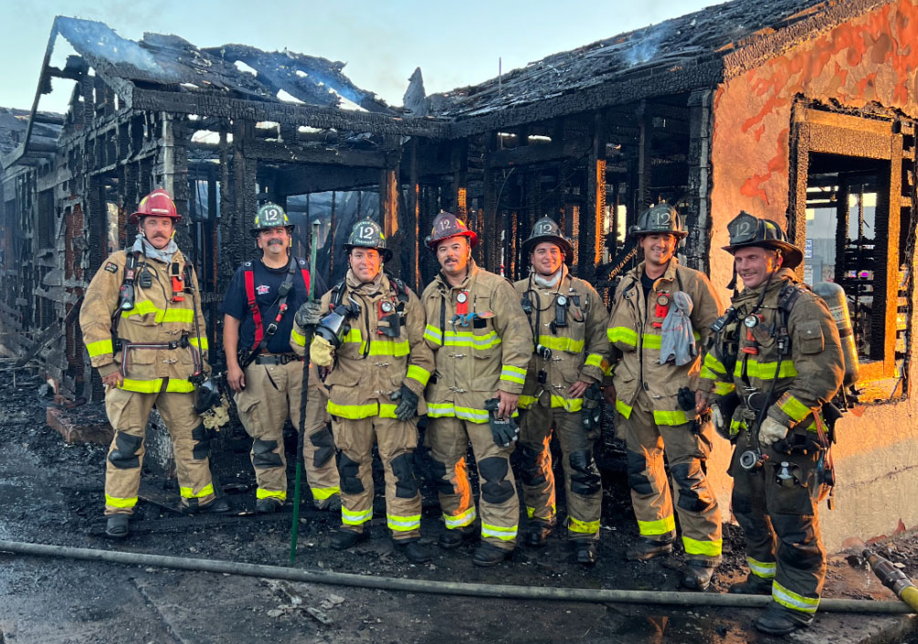 Seven firefighters wearing fire safety gear standing in front of a charred structure.