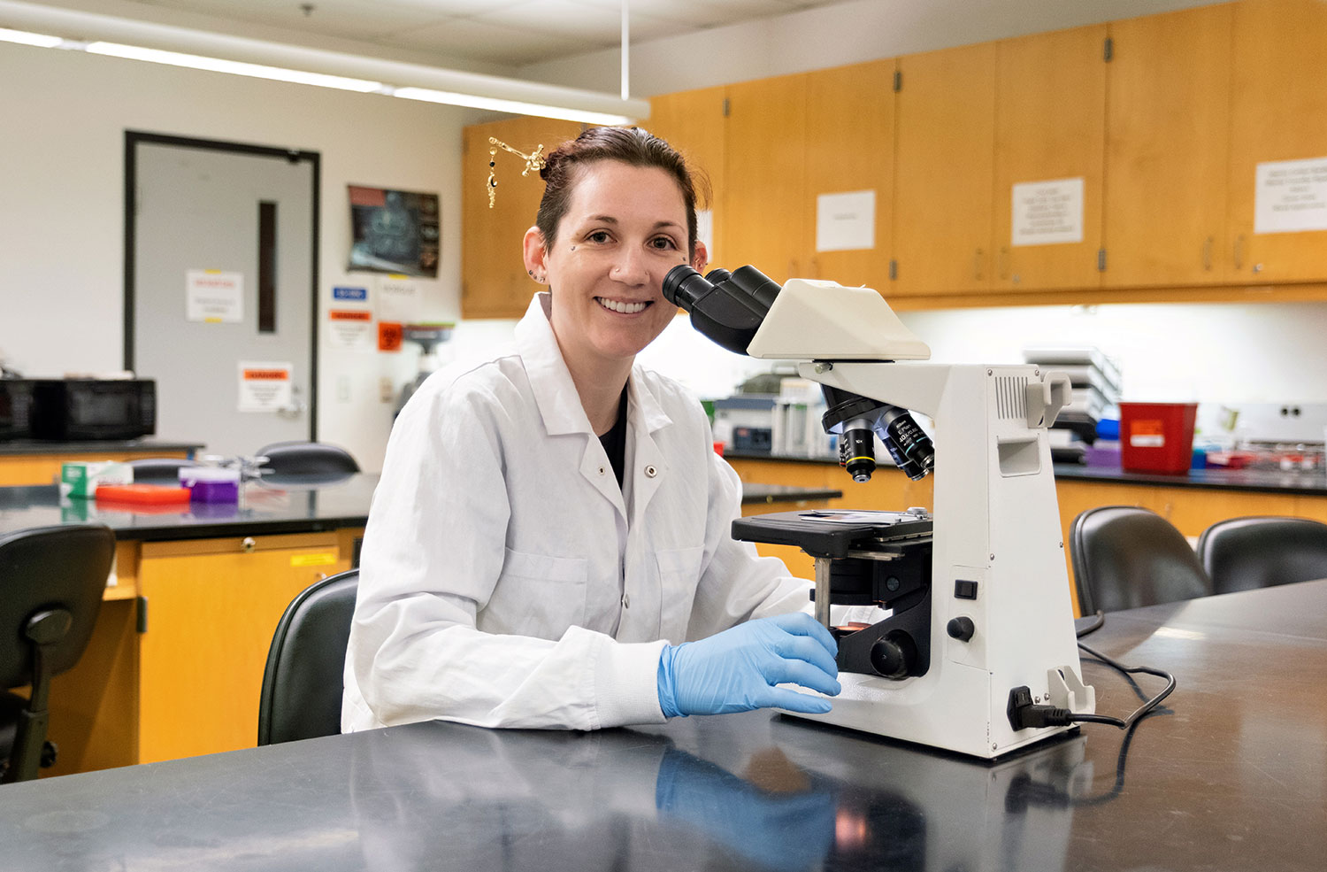Katherine Tyler sitting in front of a microscope wearing medical gloves and a white coat