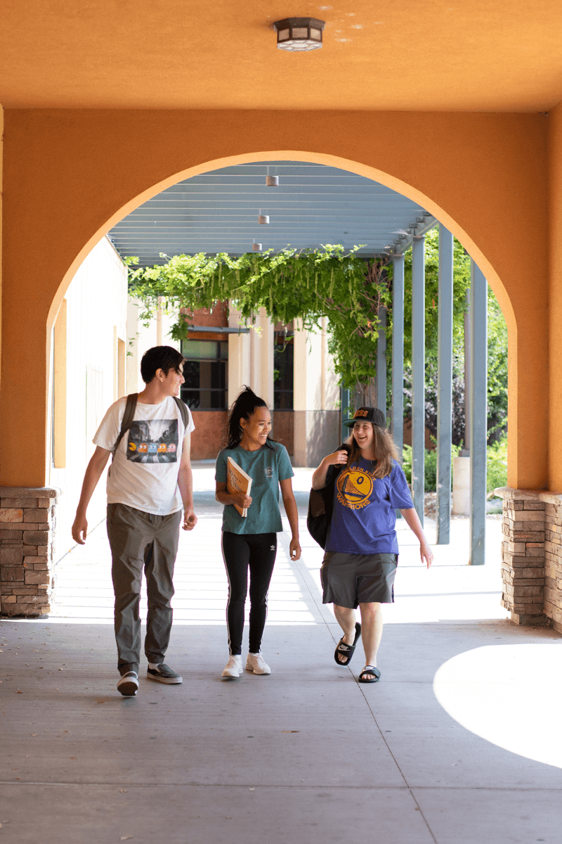 Three students walking through an archway with stone and orange walls, engaging in conversation.