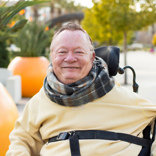 Rusty Krumm is smiling, he is sitting in a wheelchair, wearing a beige sweater and plaid scarf, outdoors with trees and large orange planters behind him.