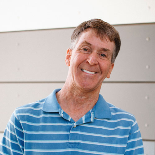 SDCCD student Gary is smiling wearing a light blue and white striped polo shirt, in front of a gray paneled wall.