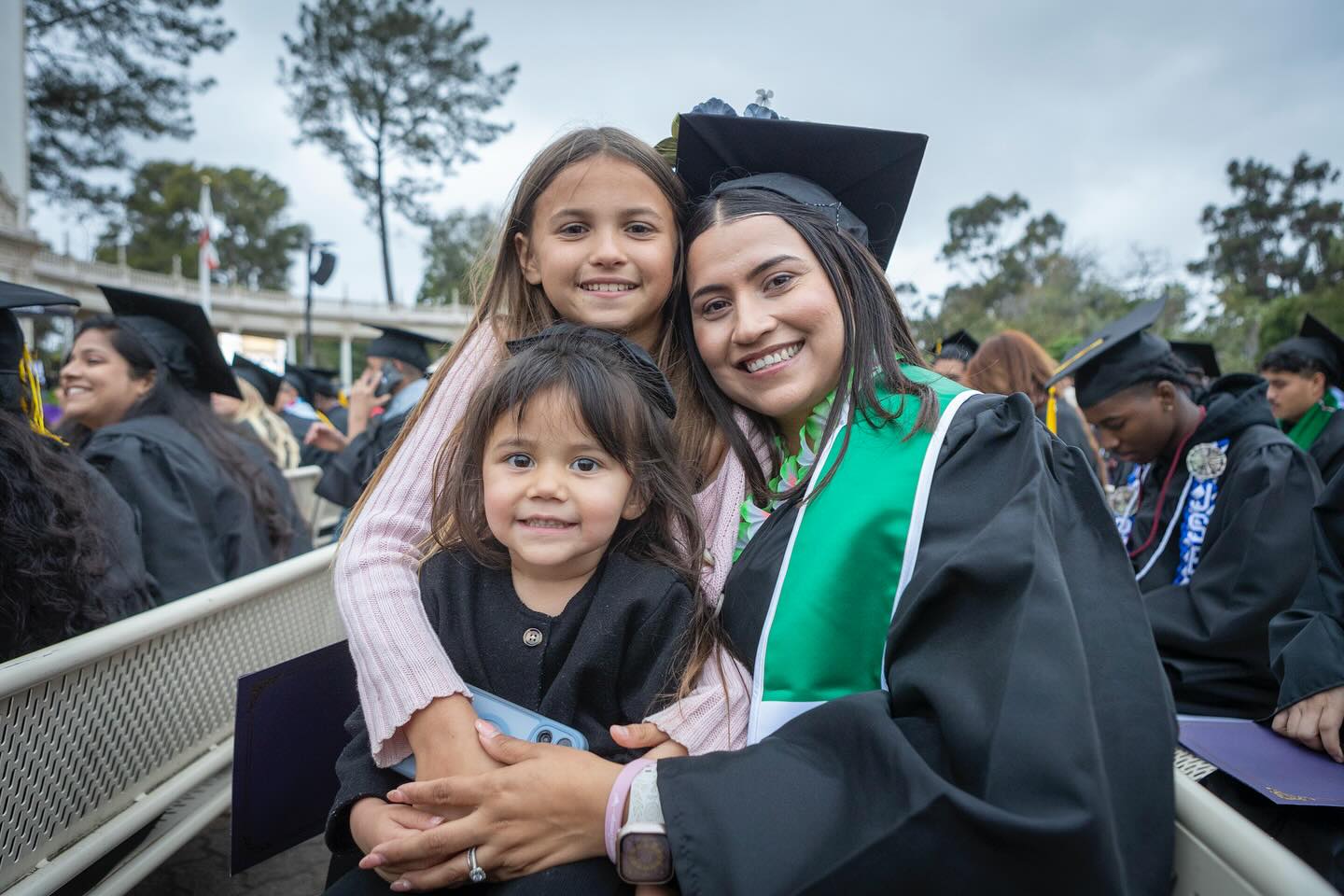 
A graduate in a black cap and gown with two young children at commencement.
