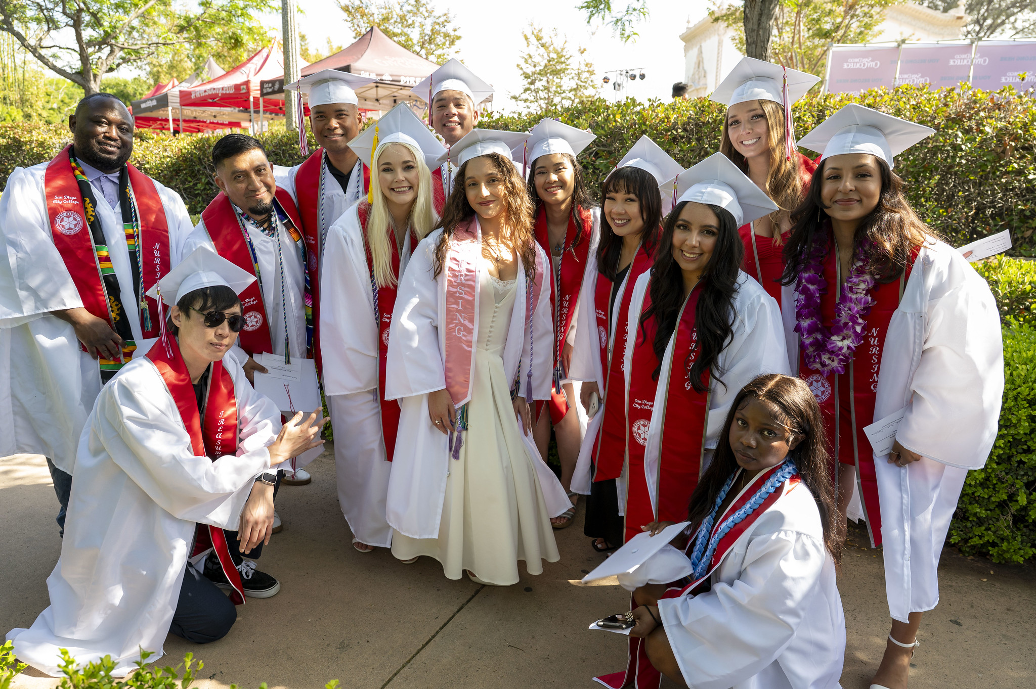 
Thirteen students in white caps and gowns with red sashes.
