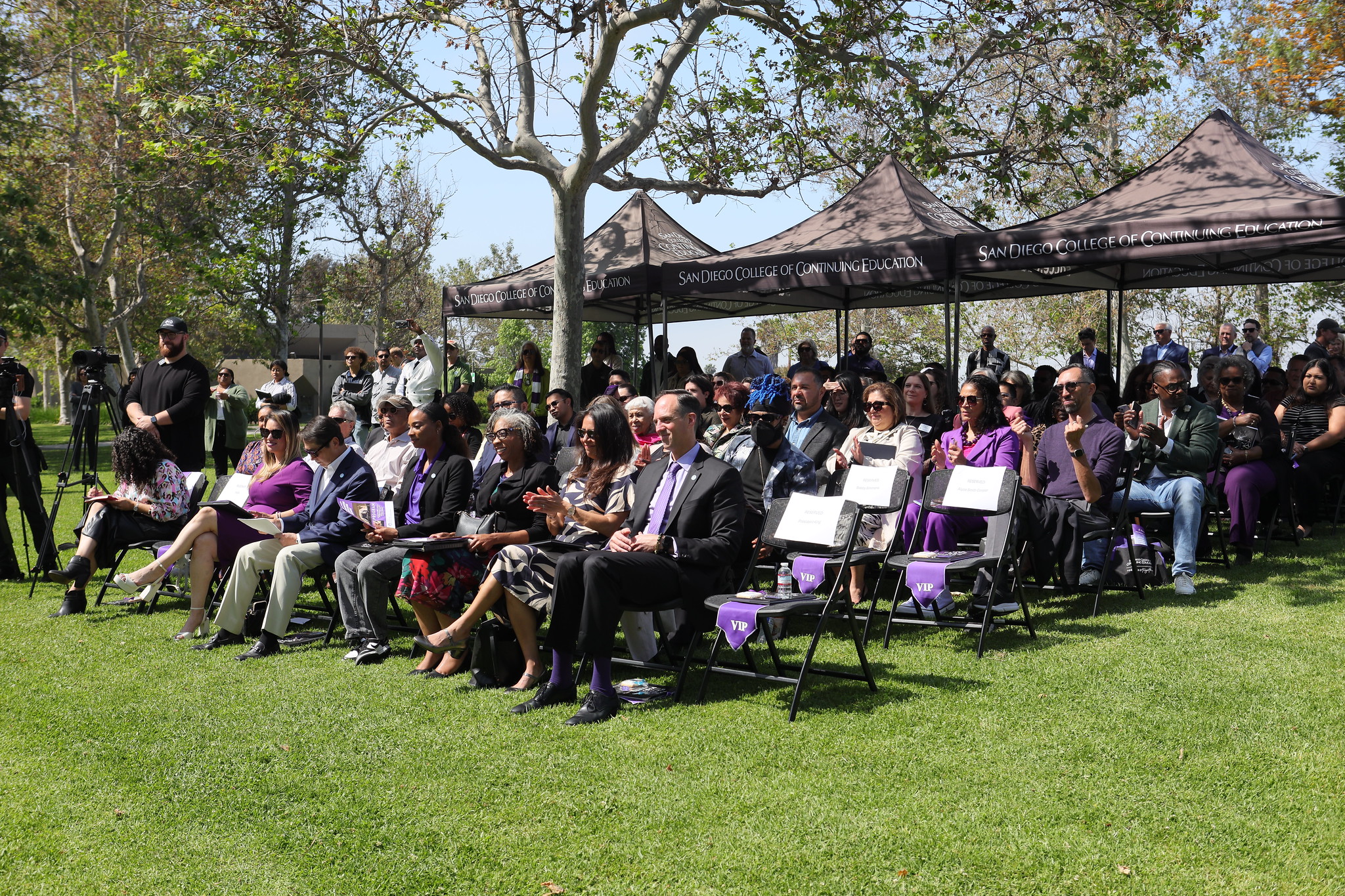 
About 75 people sit in chairs on the grass. Some stand under a shaded canopy in the back.
