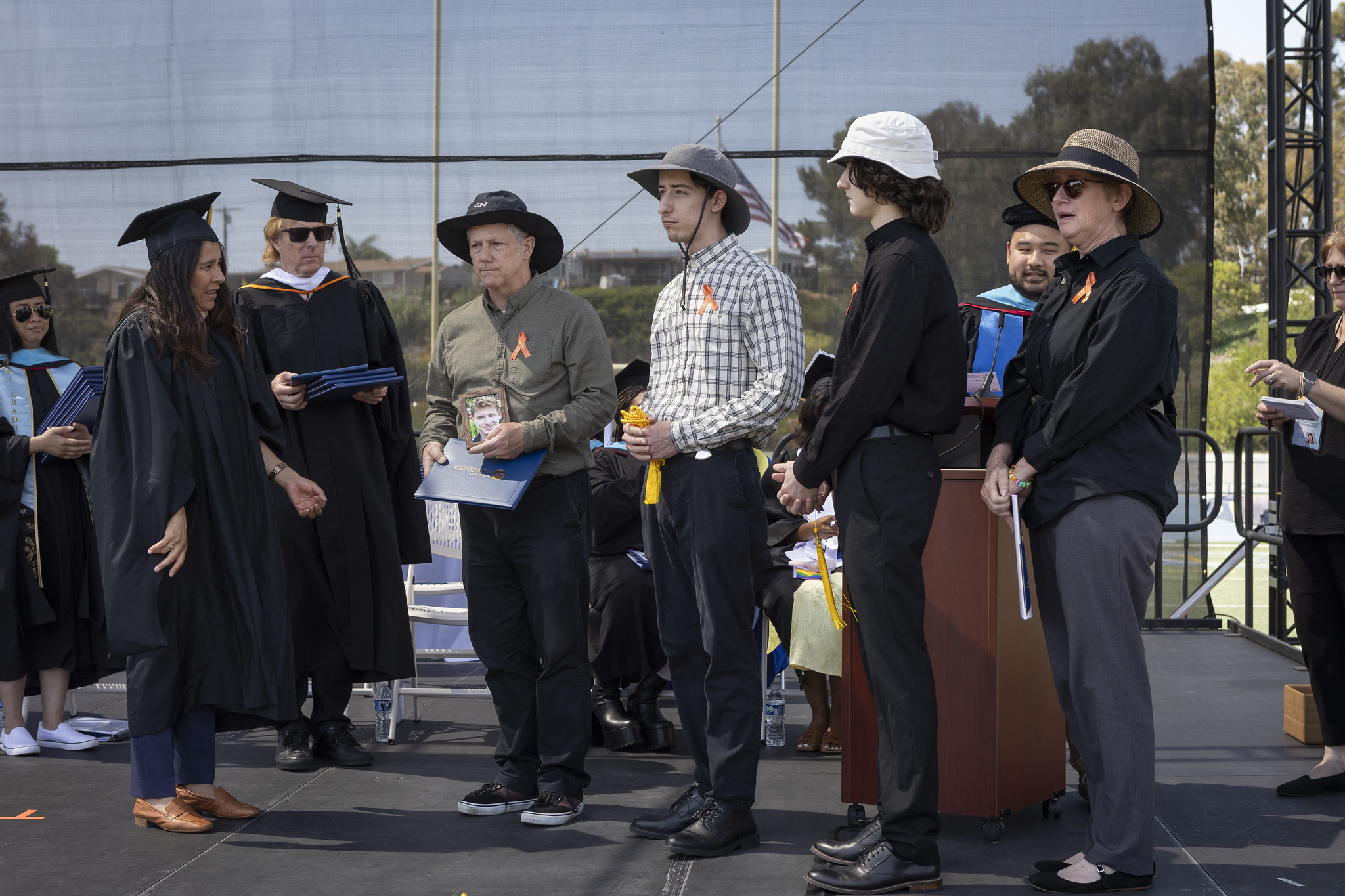 
Four family members attend commencement to receive a degree on behalf of a graduate. A man is holding the degree and a photograph of a young man.
