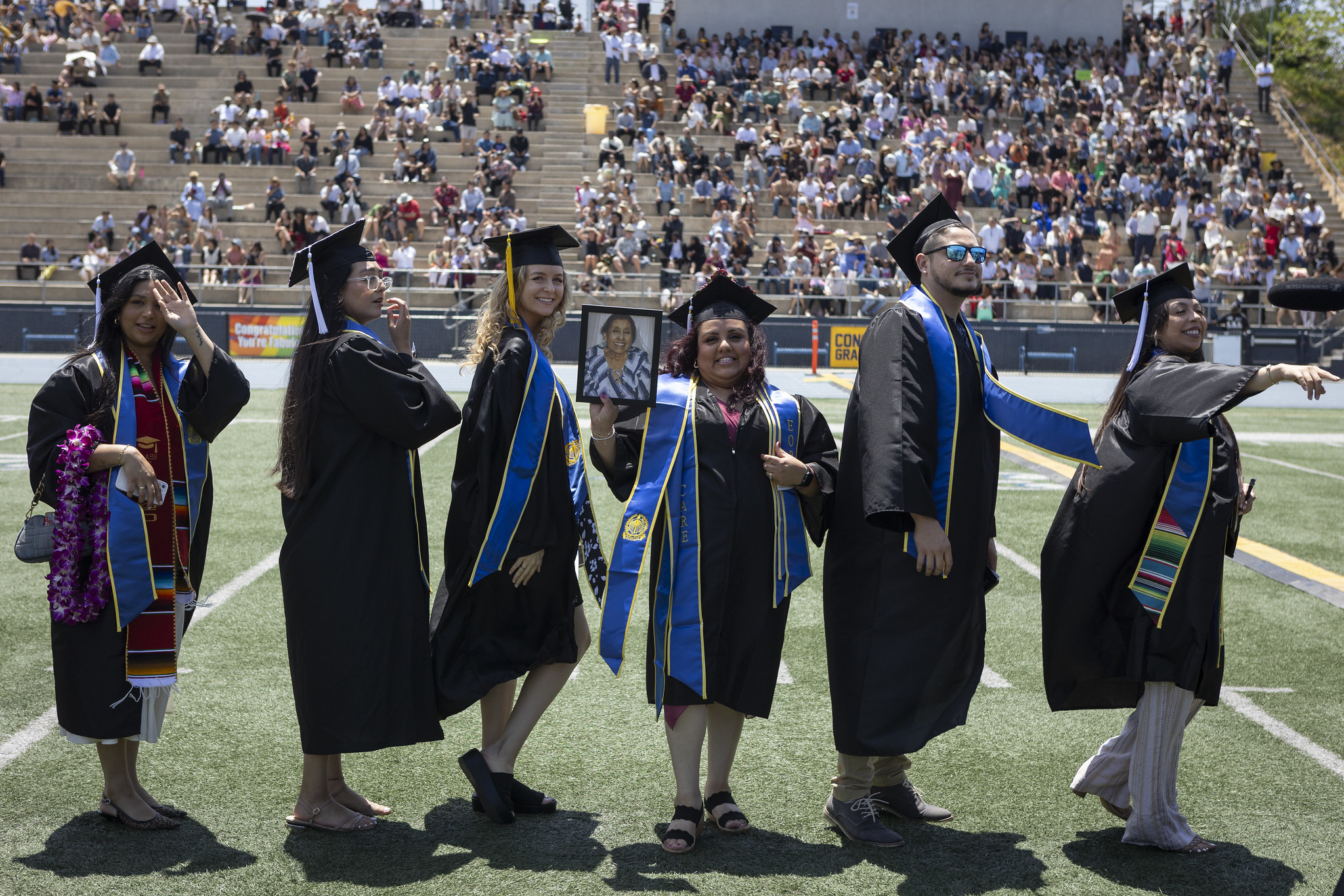 
Six graduates in a line walking to their seats and waving to the crowd.
