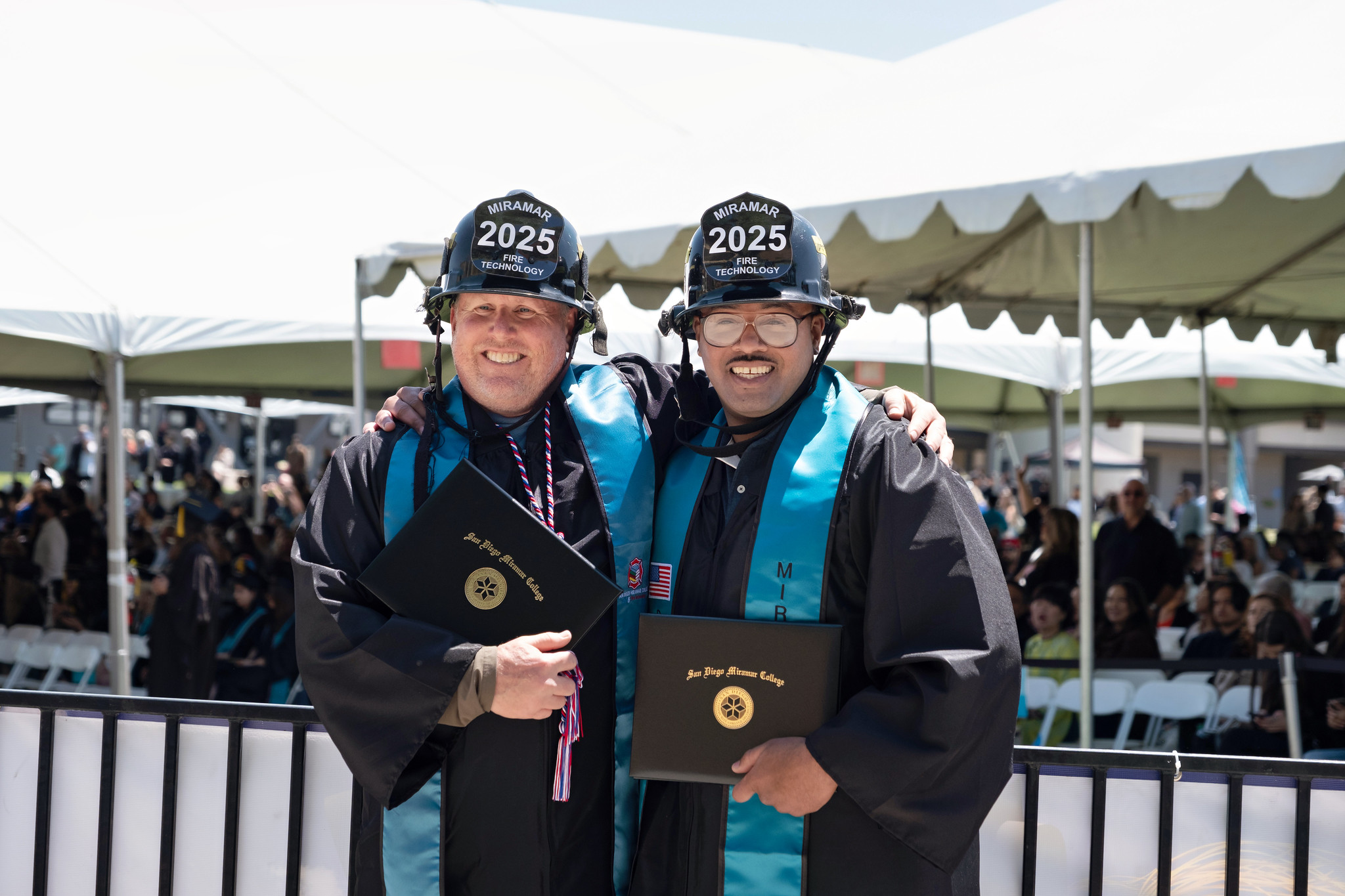 
Two fire academy students in a black fire helmets hold up their degrees.
