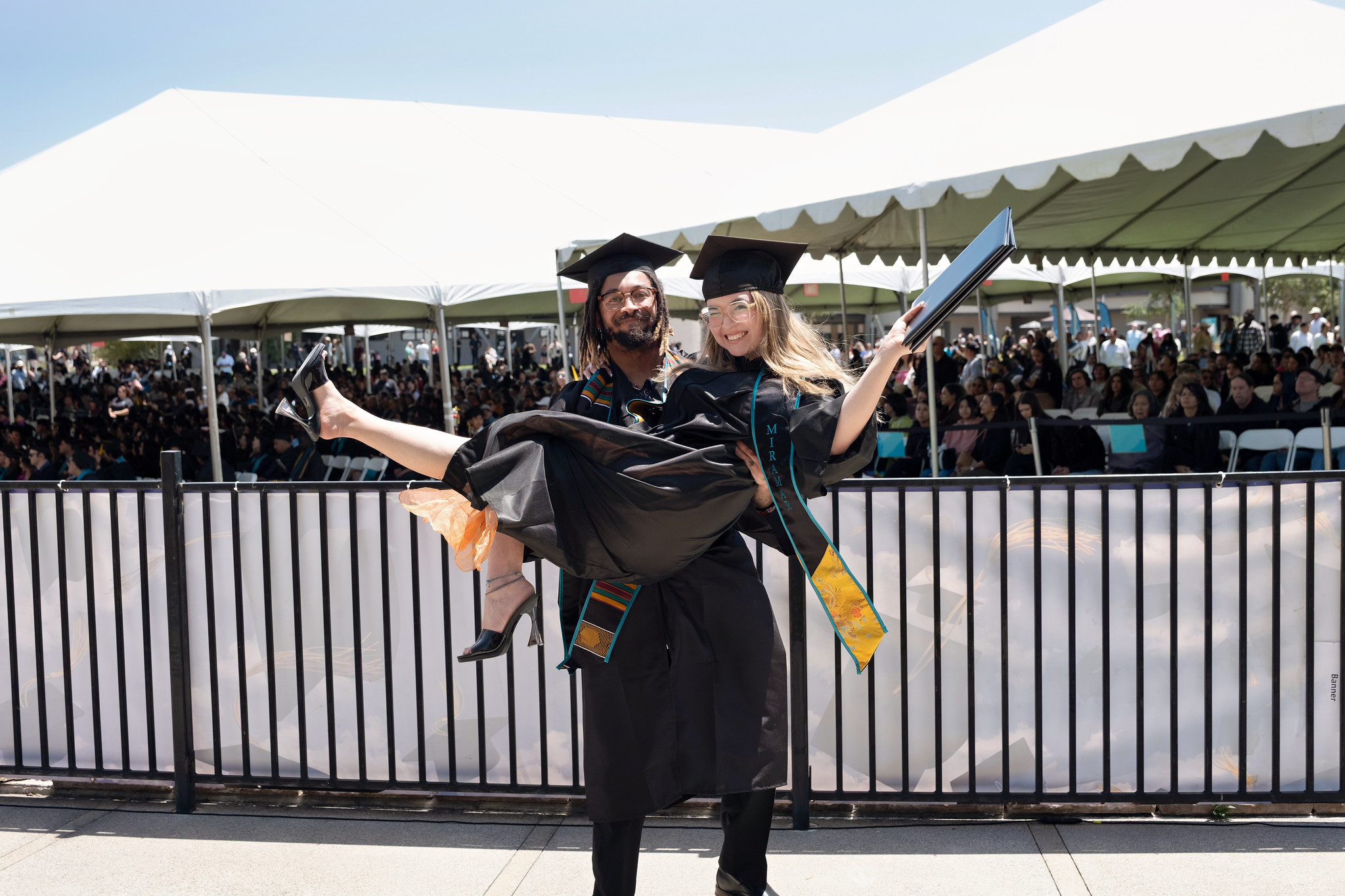 
Two graduates in black caps and gowns. The man is carrying the lady in celebration as she holds up her degree.
