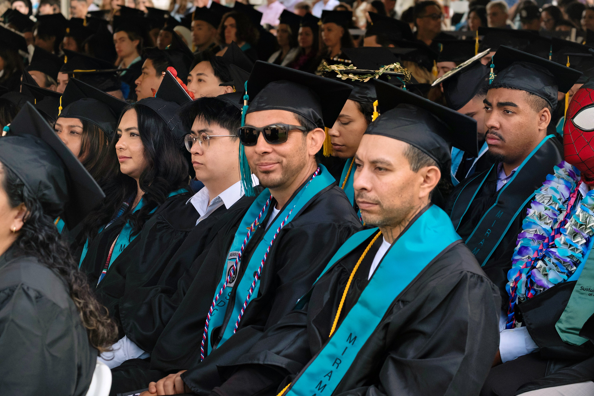 
A group of graduates in black caps and gowns with teal sashes seated during commencement.
